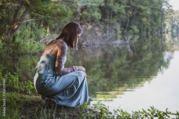 beautiful natural woman with tattoos squatting sideways on the grass on the shore of a forest lake, wearing a blue dress and looking at the water