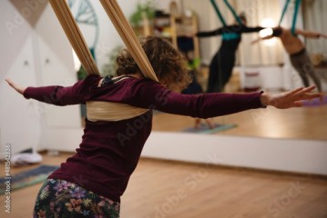 Młoda kobieta z kręconymi włosami prowadzi zajęcia z jogi na szarfach, w lustrze widać odbicie innych uczestniczek zajęć