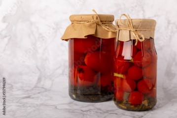 Pickled red cherry tomatoes in a glass jar on a textured marble background. Food preservation for autumn or winter. Home storage. Canning, fermentation concept. Place for text. Copy space.