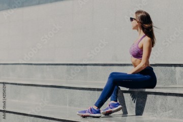 People and workout. Photo of sporty woman in purple top and sneakers, focused aside, has rest after gymanstics training, sits on stairs outdoor, wears sunglasses, likes physical activities and sport