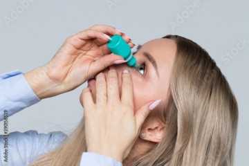 Close up of young woman applying drops in eye, grey background, isolated. Dry eyes