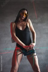 a brunette girl in a bodysuit and yellow construction glasses holds a jigsaw in her hands, stands against the background of a gray wall and a construction laser level
