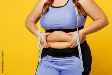 Cropped young overweight plus size big fat fit woman wear blue top warm up train hold measure tape on waist hold bely skin isolated on plain yellow background studio home gym. Workout sport concept.