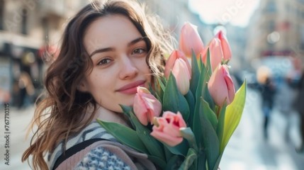 A woman is holding a bouquet of pink tulips. She is smiling and looking at the camera. The scene takes place on a city street with several other people walking around