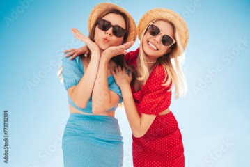 Two young beautiful smiling hipster woman in trendy summer same clothes. Carefree women in street in hats. Positive models at sunset. Cheerful and happy. Bottom view. They look at camera from above