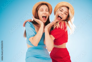 Two young beautiful smiling hipster woman in trendy summer same clothes. Carefree women in street in hats. Positive models at sunset. Cheerful and happy. Bottom view. They look at camera from above